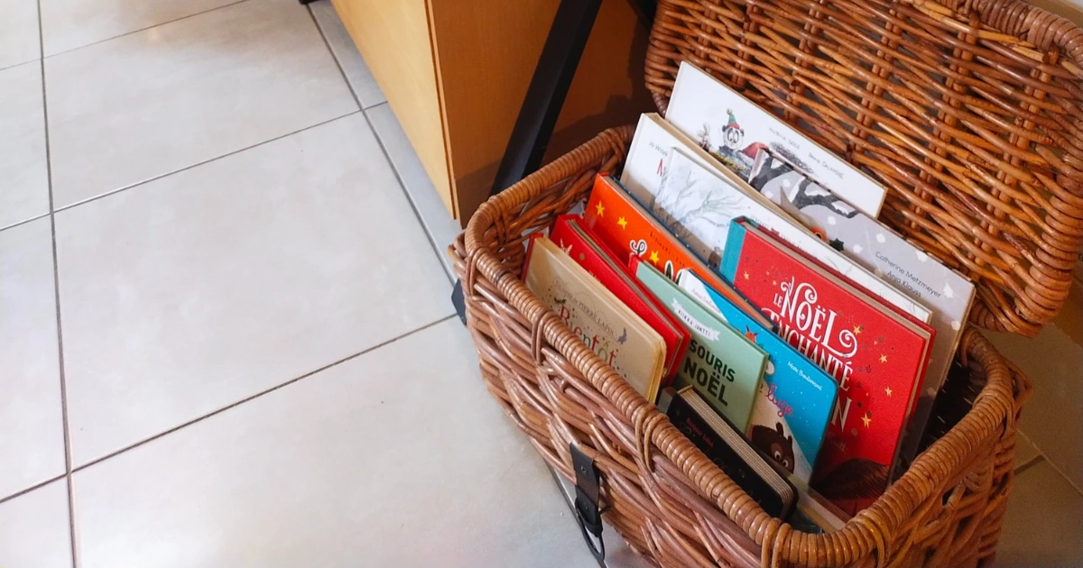 Panier en osier ouvert rempli de livres pour enfants sur le thème de Noël, posé au sol dans un salon lumineux.