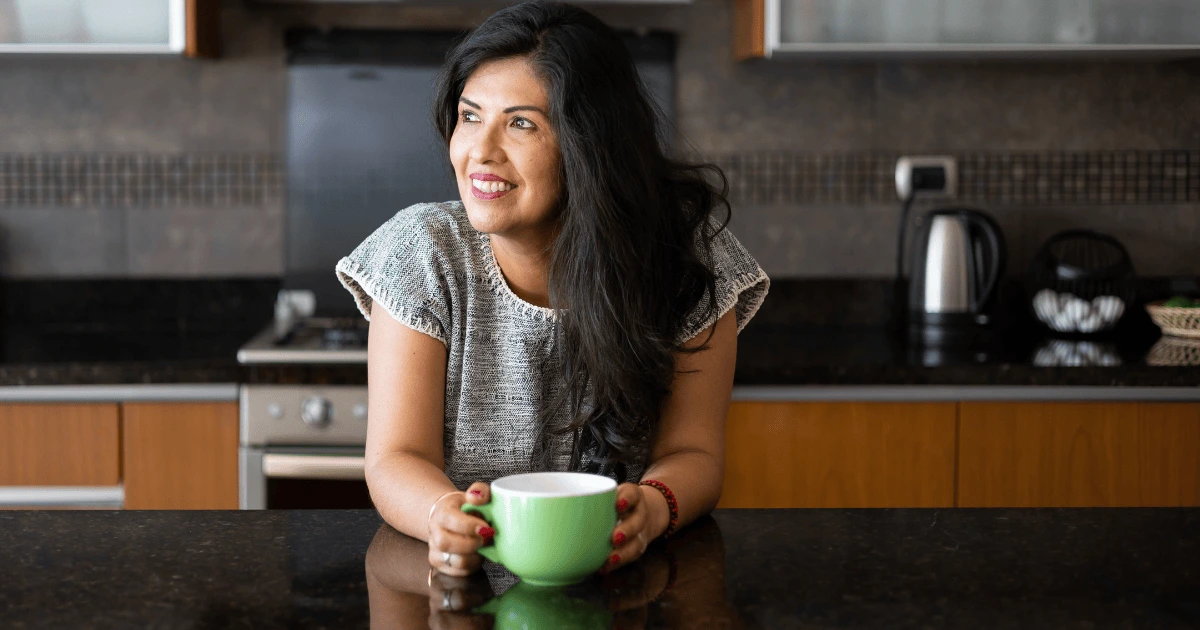 Femme appuyée sur un plan de travail dans une cuisine, tenant une tasse de café, illustrant un quotidien plus facile et agréable à vivre.