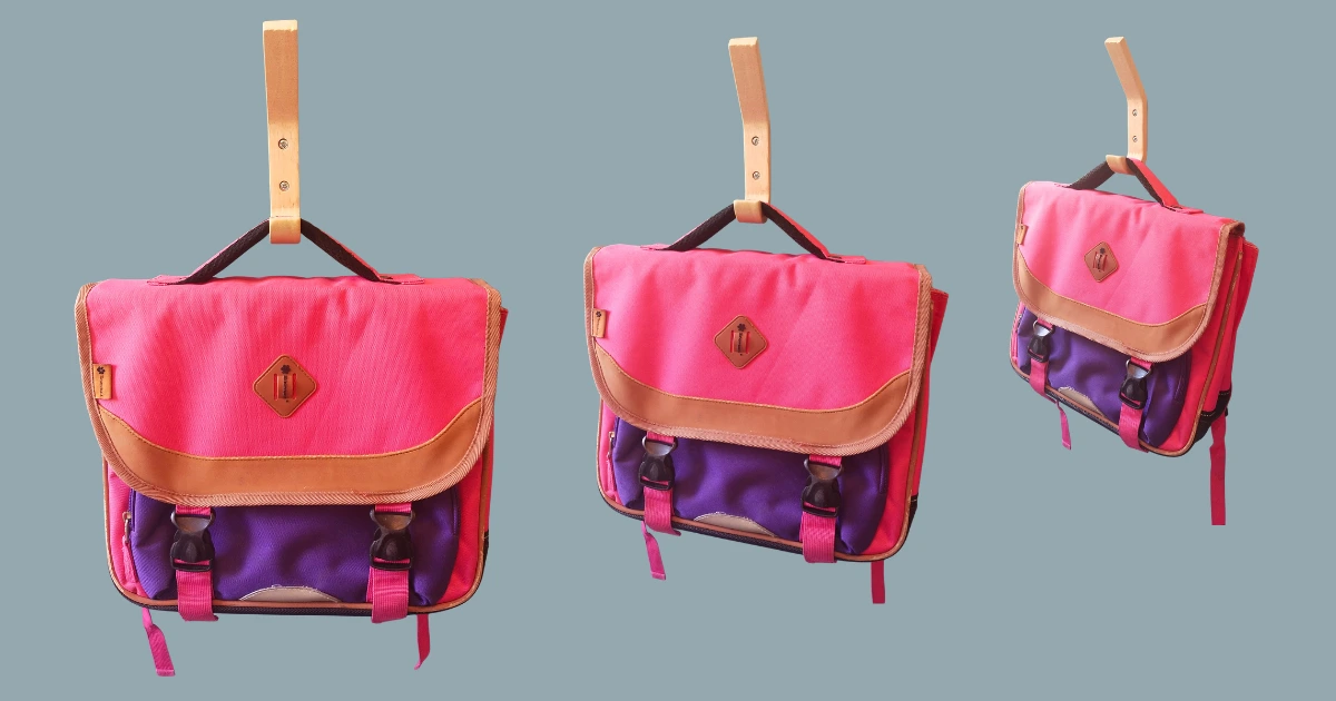 Visuel montrant trois cartables d'écolier roses et violets suspendus à des patères murales, présentés dans différentes tailles. L'image illustre l'article sur le tri des affaires scolaires dès le début des vacances d'été afin de préparer la rentrée, vérifier le matériel réutilisable et anticiper les achats scolaires.